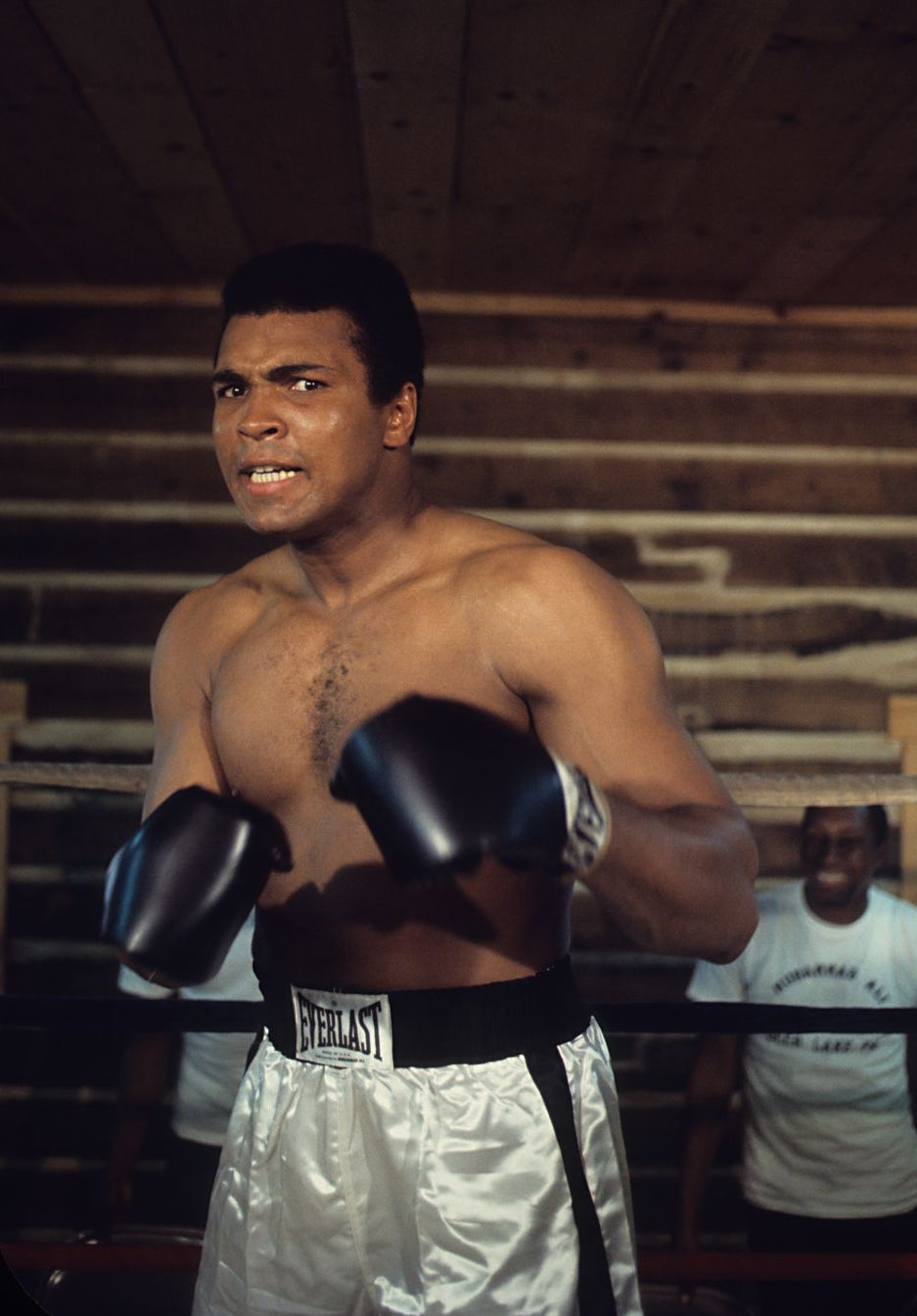 Muhammad Ali in a boxing ring wearing black Everlast boxing gloves and white satin shorts, with a wooden boxing gym background.